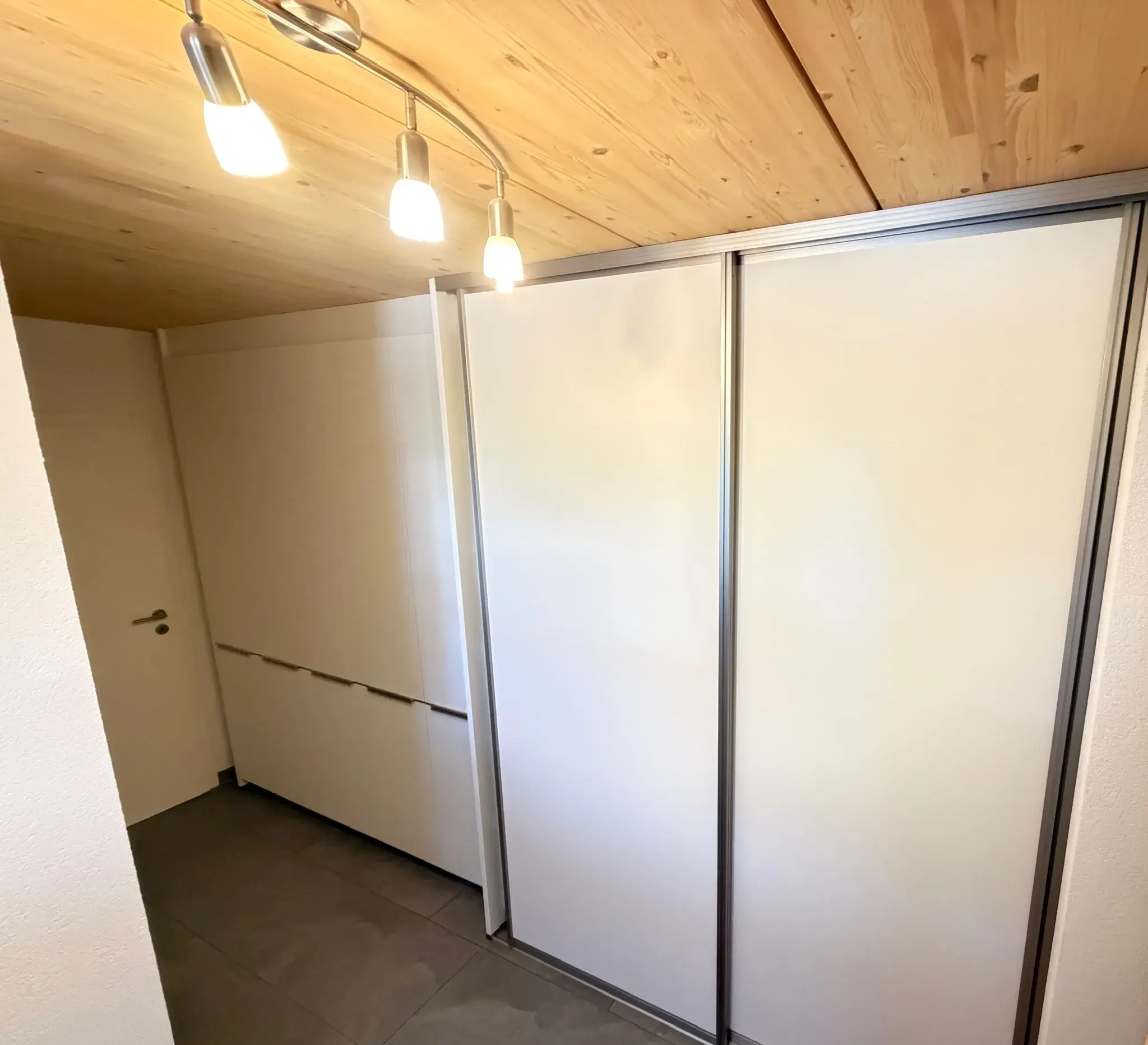 Flur mit weißem Schiebetürenschrank und Holzdecke, beleuchtet von drei Lampen.