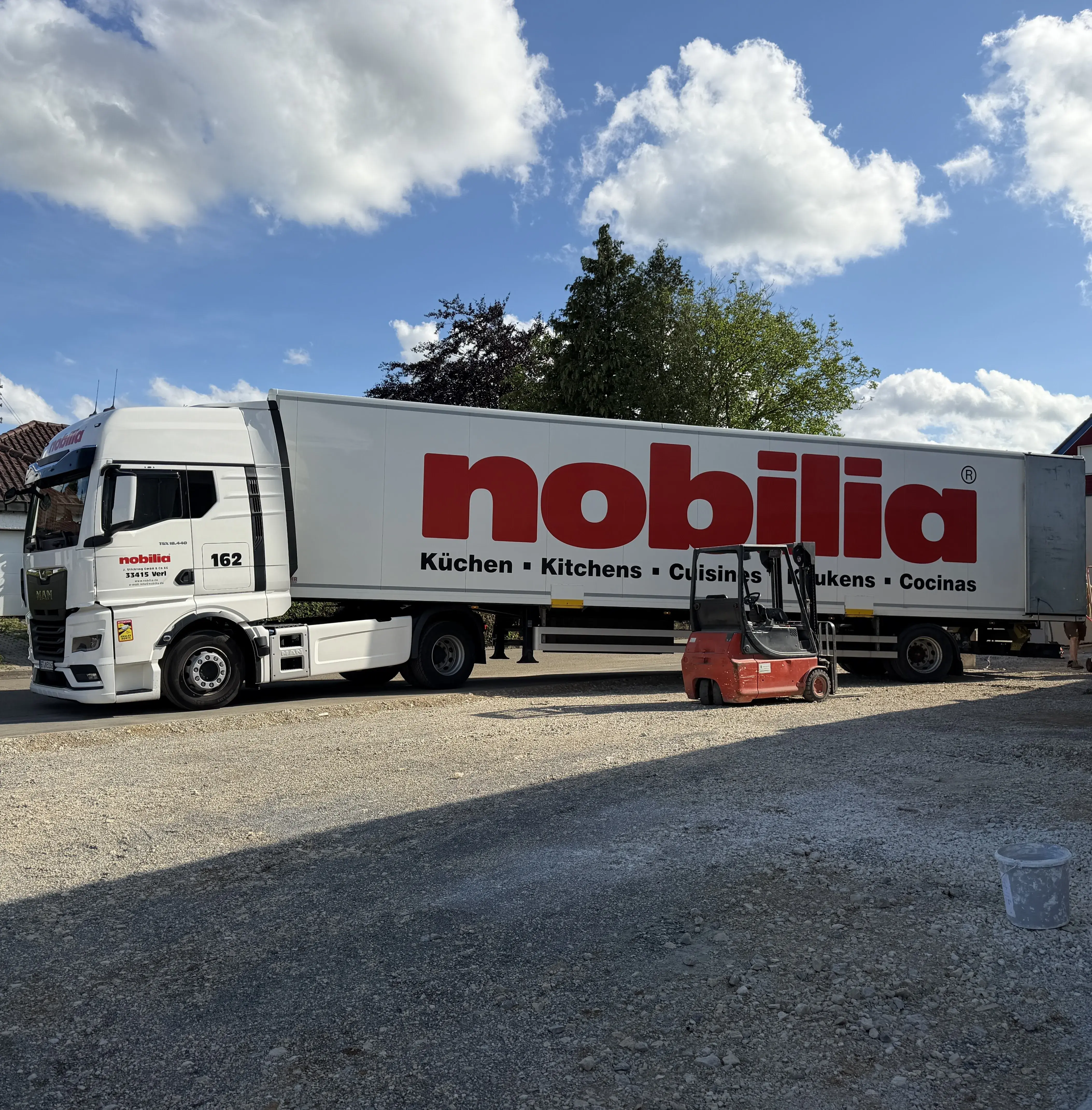 LKW weiß mit roter Aufschrift, Stapler, Schotter Parkplatz, Wolken, Blauer Himmel
